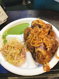 A plate of grilled meatballs topped with sauteed onions and a vibrant orange sauce. The dish is accompanied by a serving of finely shredded coleslaw with onions and thin slices of cabbage, placed next to a dip of green chutney.