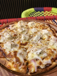 A freshly baked pizza topped with melted cheese, chunks of feta, and sprinkled with herbs. The crust is golden brown, and there are visible onion slices scattered over the top. The pizza is placed on a wooden tray with a colorful woven background featuring patterns in red, green, and blue.