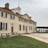 A historical colonial-style mansion with a symmetrical facade, featuring large shuttered windows and a prominent cupola. The building is surrounded by a well-maintained lawn and a paved courtyard. The architecture reflects classic elements with a beige exterior and a red brick roof.