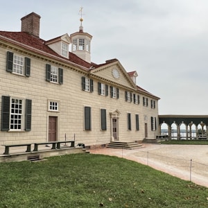 A historical colonial-style mansion with a symmetrical facade, featuring large shuttered windows and a prominent cupola. The building is surrounded by a well-maintained lawn and a paved courtyard. The architecture reflects classic elements with a beige exterior and a red brick roof.