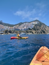 A person is kayaking on calm blue waters near a coastal area with hillside buildings visible in the background. The kayaker wears a life vest and helmet, paddling in a brightly colored kayak. The sun is shining, and the scene depicts a serene outdoor setting.