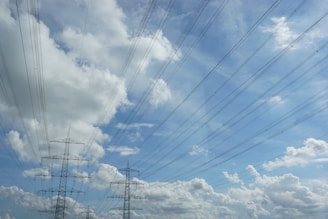 High-voltage power lines stretch across a blue sky filled with fluffy white clouds. Metal lattice towers support the wires, which fan out in various directions.