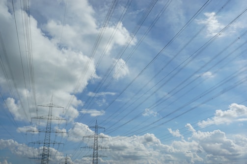 High-voltage power lines stretch across a blue sky filled with fluffy white clouds. Metal lattice towers support the wires, which fan out in various directions.