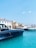 A marina with several luxury yachts docked in turquoise blue water. The sky is clear with minimal clouds and there are buildings with terracotta roofs in the background.