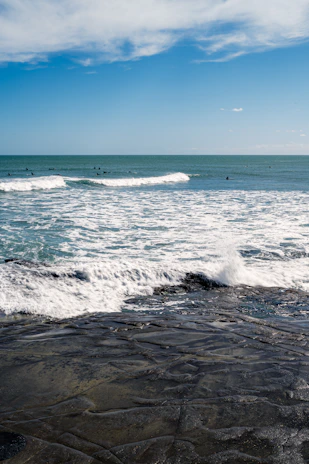 Surfers catching waves under a bright blue sky, with the shoreline visible in the distance.