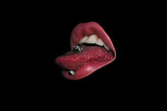A mouth with a tongue piercing, featuring a silver ball piercing, against a black background. The lips are prominently displayed and have a glossy appearance, with visible teeth.