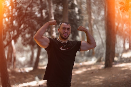 A man stands in a forest flexing his biceps, wearing a black T-shirt with a white logo. He is posing playfully with his tongue out, surrounded by trees and soft sunlight filtering through foliage.