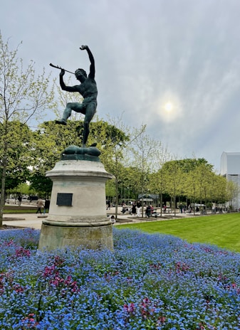 A bronze statue of a man playing a flute is prominently displayed atop a pedestal. The foreground features a bed of vibrant blue and pink flowers, and the scene is set in a park with a backdrop of trees. The sky is overcast with a sun peeking through the clouds, creating a calm ambiance. People can be seen strolling in the background.