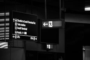 The image features airport signage with directions to different locations including the shuttle for the South Terminal, train station, South Terminal, hotels, and lifts. The signs are hanging from the ceiling and are displayed in white lettering on a dark background. An emergency exit sign with an arrow pointing left is also visible.