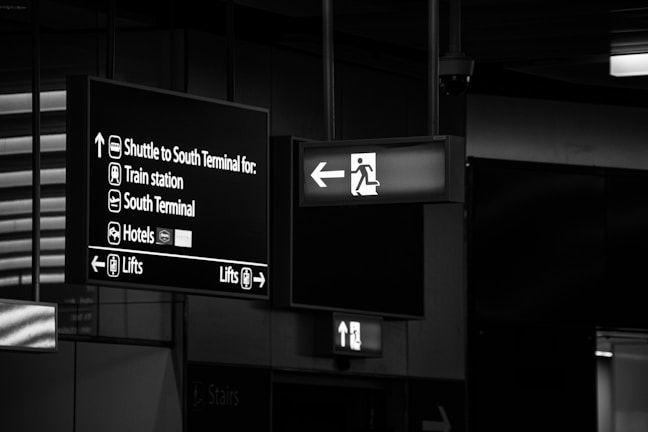 The image features airport signage with directions to different locations including the shuttle for the South Terminal, train station, South Terminal, hotels, and lifts. The signs are hanging from the ceiling and are displayed in white lettering on a dark background. An emergency exit sign with an arrow pointing left is also visible.