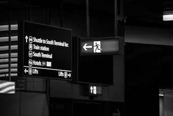 The image features airport signage with directions to different locations including the shuttle for the South Terminal, train station, South Terminal, hotels, and lifts. The signs are hanging from the ceiling and are displayed in white lettering on a dark background. An emergency exit sign with an arrow pointing left is also visible.