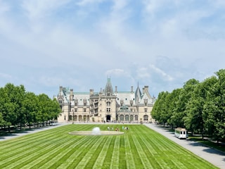An elegant, large estate with intricate architecture, featuring numerous spires and chimneys. A manicured lawn with symmetrical stripes leads to a fountain in front of the building. Lush green trees line both sides of the pathway, and there is a tram on a paved road to the right.