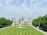 An elegant, large estate with intricate architecture, featuring numerous spires and chimneys. A manicured lawn with symmetrical stripes leads to a fountain in front of the building. Lush green trees line both sides of the pathway, and there is a tram on a paved road to the right.
