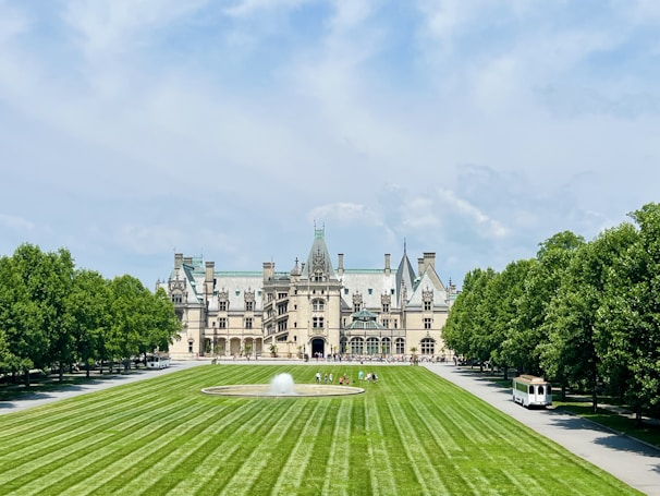 An elegant, large estate with intricate architecture, featuring numerous spires and chimneys. A manicured lawn with symmetrical stripes leads to a fountain in front of the building. Lush green trees line both sides of the pathway, and there is a tram on a paved road to the right.