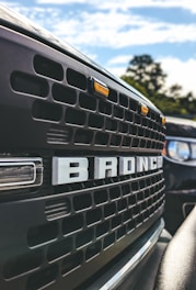 Close-up view of a black vehicle's front grille with a prominent 'BRONCO' emblem. The sky is partly cloudy with some trees visible in the background.