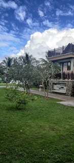 A lush green garden with a well-maintained grassy lawn is seen under a bright blue sky. The garden includes a few small trees and shrubs. In the background, a two-story house with a stone facade and wooden balcony railings can be observed. The sky is partly cloudy with large, fluffy clouds.