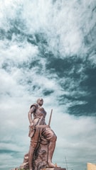 A prominent statue of a warrior stands against a dramatic cloudy sky. The figure is depicted in a dynamic pose, holding a sword and wearing traditional attire. The statue is mounted on a large rock base, situated in an open area.