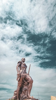 A prominent statue of a warrior stands against a dramatic cloudy sky. The figure is depicted in a dynamic pose, holding a sword and wearing traditional attire. The statue is mounted on a large rock base, situated in an open area.
