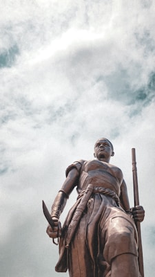 A stately bronze statue of a warrior stands against a backdrop of cloudy skies. The figure is adorned in traditional attire and holds a weapon in each hand, exuding strength and determination.