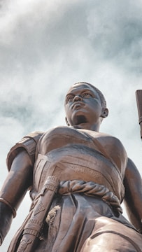 A majestic bronze statue of a warrior looking upwards, appearing powerful and determined. The figure is clad in detailed traditional attire, including a sash and arm bands, with a confident and resolute expression. The angle of the shot enhances the grandeur and imposing nature of the statue, with a backdrop of a clouded sky.
