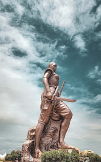 A large statue of a warrior holding a sword stands prominently against a dramatic sky. The statue, detailed in its depiction of armor and weaponry, is positioned on a rocky base surrounded by a few people and greenery. The sky is filled with clouds, adding depth to the scene.