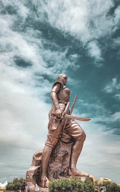 A large statue of a warrior holding a sword stands prominently against a dramatic sky. The statue, detailed in its depiction of armor and weaponry, is positioned on a rocky base surrounded by a few people and greenery. The sky is filled with clouds, adding depth to the scene.