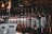 A row of glass bottles with pumps, each labeled with different flavor names like 'Maple', 'Bubble Gum', and 'Green Mint'. Dimly lit setting enhances the ambiance with a variety of labels visible on the bottles. There are also menu cards in front of the bottles displaying a selection of items.