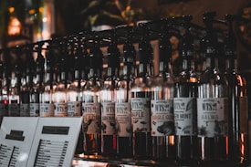 A row of glass bottles with pumps, each labeled with different flavor names like 'Maple', 'Bubble Gum', and 'Green Mint'. Dimly lit setting enhances the ambiance with a variety of labels visible on the bottles. There are also menu cards in front of the bottles displaying a selection of items.