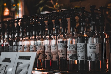 A row of glass bottles with pumps, each labeled with different flavor names like 'Maple', 'Bubble Gum', and 'Green Mint'. Dimly lit setting enhances the ambiance with a variety of labels visible on the bottles. There are also menu cards in front of the bottles displaying a selection of items.