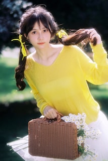 A person with long hair tied in braids adorned with yellow ribbons holds them out to the side. They are wearing a bright yellow sweater and stand holding a wicker basket filled with white daisies. The background is a natural setting with green foliage.