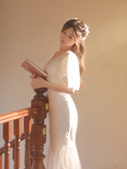 A woman in a light-colored, flowing dress is holding a book in a serene and contemplative pose next to a wooden staircase. Soft natural light illuminates her and creates a warm ambiance.