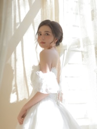A young woman is standing in soft natural light, wearing an elegant off-the-shoulder white dress. Her hair is styled in a loose updo, and she gazes directly at the camera with a serene expression. The backdrop consists of sheer curtains with sunlight streaming through, casting gentle shadows.