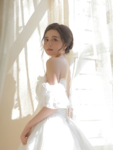 A young woman is standing in soft natural light, wearing an elegant off-the-shoulder white dress. Her hair is styled in a loose updo, and she gazes directly at the camera with a serene expression. The backdrop consists of sheer curtains with sunlight streaming through, casting gentle shadows.