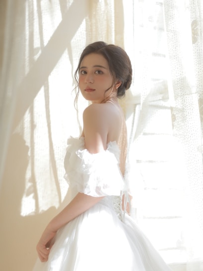 A softly lit studio photo of a model wearing a chic, flowing dress from the collection, standing beside a window with gentle natural light.