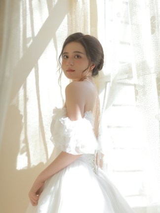 A young woman is standing in soft natural light, wearing an elegant off-the-shoulder white dress. Her hair is styled in a loose updo, and she gazes directly at the camera with a serene expression. The backdrop consists of sheer curtains with sunlight streaming through, casting gentle shadows.