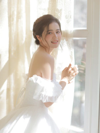 A woman in a light linen dress enjoying morning sunlight in a minimalist, neutral-toned room.
