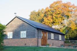 A cozy wooden cabin with a brick foundation is set against the backdrop of vibrant autumn trees with colorful foliage. There is a chimney emitting smoke, suggesting warmth and comfort inside. Several potted plants are arranged in front of the building, enhancing the rustic charm. The grass in the foreground is lush and green.
