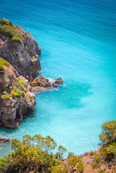 Turquoise blue ocean waters meet rugged rocky cliffs with patches of green vegetation. Sunlight glistens on the water's surface, creating a serene and inviting atmosphere.