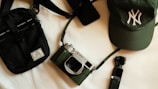 A collection of items including a black crossbody bag, a green cap with an embroidered logo, a vintage-style camera with a black and silver finish, a small black handheld device, and a smartphone. These objects are arranged on a light surface, creating an aesthetically pleasing composition.