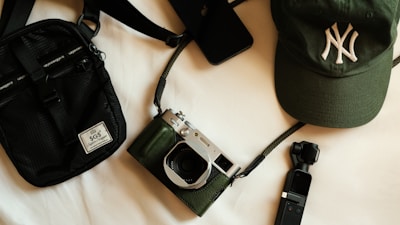 A collection of items including a black crossbody bag, a green cap with an embroidered logo, a vintage-style camera with a black and silver finish, a small black handheld device, and a smartphone. These objects are arranged on a light surface, creating an aesthetically pleasing composition.
