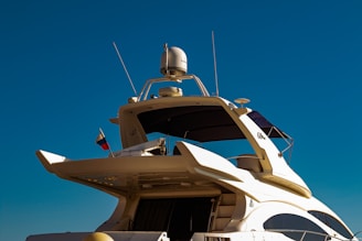 A luxury yacht with a sleek, modern design features multiple decks and advanced equipment visible on top. A small flag flutters near the rear, and the sky is clear and blue.