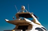 A luxury yacht with a sleek, modern design features multiple decks and advanced equipment visible on top. A small flag flutters near the rear, and the sky is clear and blue.