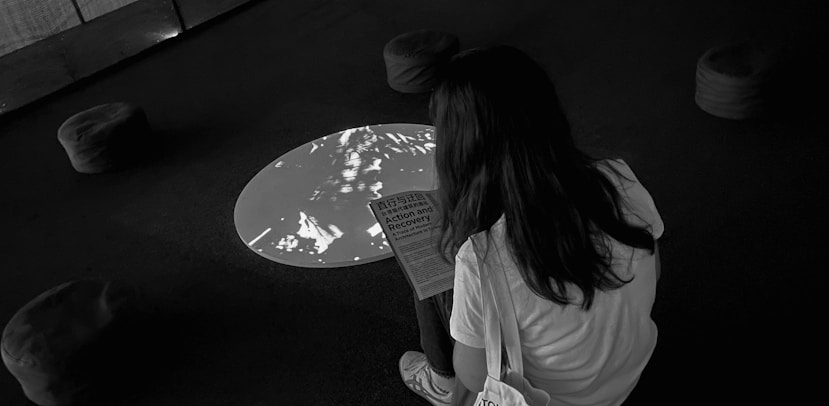 A person with long hair is sitting on a stool in a dark room, reading a document titled 'Action and Recovery'. The room features a circular light or projection on the floor and several cushioned stools scattered around.