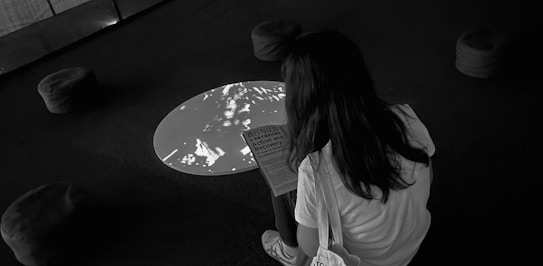 A person with long hair is sitting on a stool in a dark room, reading a document titled 'Action and Recovery'. The room features a circular light or projection on the floor and several cushioned stools scattered around.