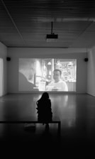 A person sits in a dark room on a bench, watching a black and white film projected on a large screen. The room is spacious with a high ceiling and two rectangular windows with curtains on either side of the screen. The film being shown features a scene with two people in what appears to be a framed photos-filled setting, and there are subtitles present.