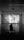 A person sits in a dark room on a bench, watching a black and white film projected on a large screen. The room is spacious with a high ceiling and two rectangular windows with curtains on either side of the screen. The film being shown features a scene with two people in what appears to be a framed photos-filled setting, and there are subtitles present.