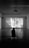 A person sits in a dark room on a bench, watching a black and white film projected on a large screen. The room is spacious with a high ceiling and two rectangular windows with curtains on either side of the screen. The film being shown features a scene with two people in what appears to be a framed photos-filled setting, and there are subtitles present.