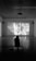 A person sits in a dark room on a bench, watching a black and white film projected on a large screen. The room is spacious with a high ceiling and two rectangular windows with curtains on either side of the screen. The film being shown features a scene with two people in what appears to be a framed photos-filled setting, and there are subtitles present.