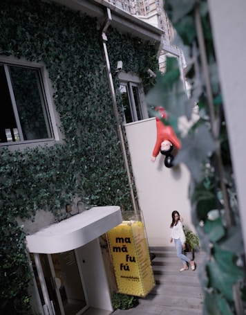 An outdoor area with walls covered in dense green ivy. A woman in casual attire ascends a flight of stairs beside a large yellow display filled with rubber balls and the text 'mam&aacute; fuf&uacute;'. Above her, a whimsical sculpture of a child in a red outfit is mounted to the wall. A modern building with large windows is partially visible in the background.