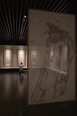 A dimly lit art gallery with large framed artworks on the walls. In the foreground, a detailed sketch of a person wearing traditional attire is partially visible. In the background, a visitor sits on a bench, observing the paintings.